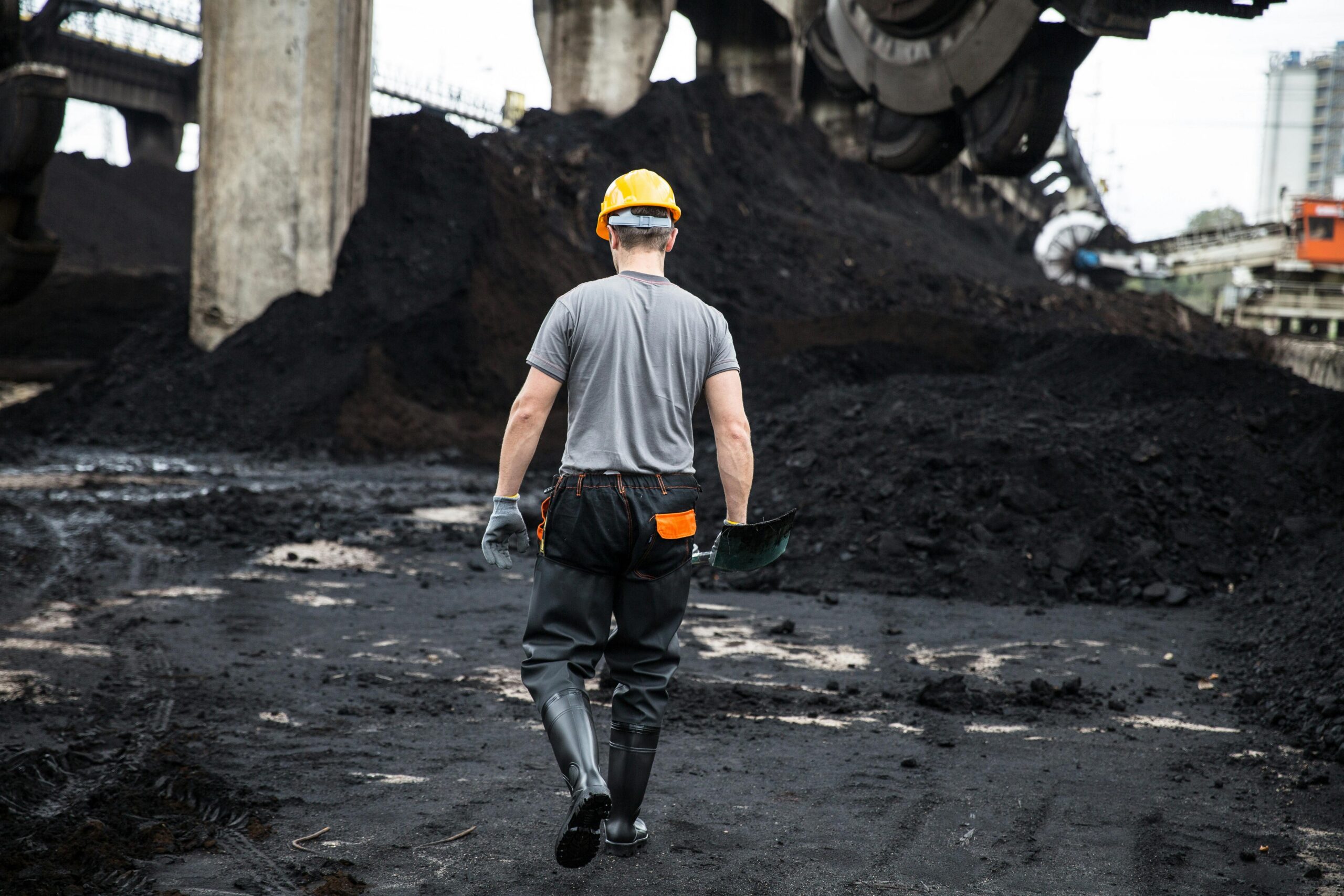 A construction worker with a hard hat walks through an industrial site filled with dirt and machinery.